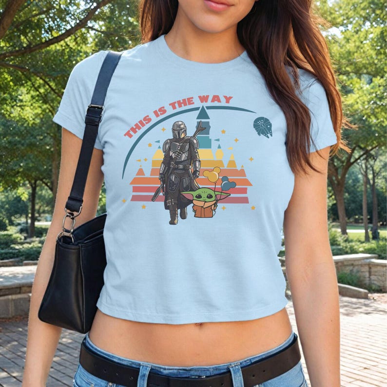 May include: Light blue crop top with a graphic featuring a Mandalorian character and Grogu, with the text "THIS IS THE WAY". The design includes a castle, a sunset, and a spaceship.