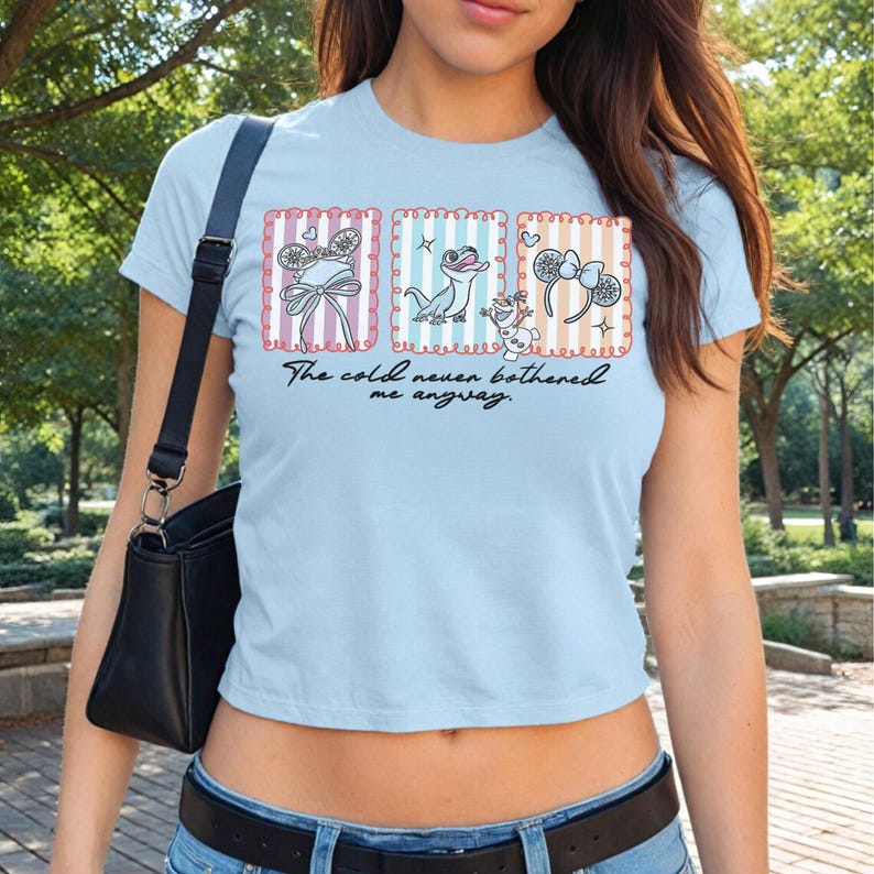 May include: Pale blue t-shirt with three framed illustrations of Frozen characters. The text "The cold never bothered me anyway" is printed on the shirt. The illustrations include a bow, a lizard, and a snowman.