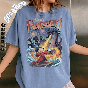 May include: Blue t-shirt featuring a colorful graphic of Mickey Mouse in a red robe, facing a dragon breathing fire. The text "Disney's Fantasmic!" is above the characters. Fireworks and other characters are in the background.