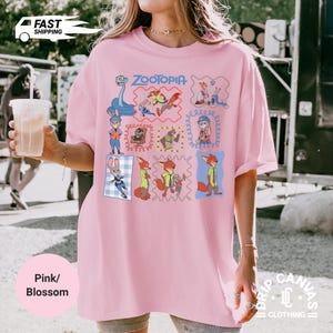 May include: Pink t-shirt with a Zootopia graphic featuring various characters in framed illustrations. The shirt has a crew neck and short sleeves. The text "Zootopia" is in blue script above the character illustrations.