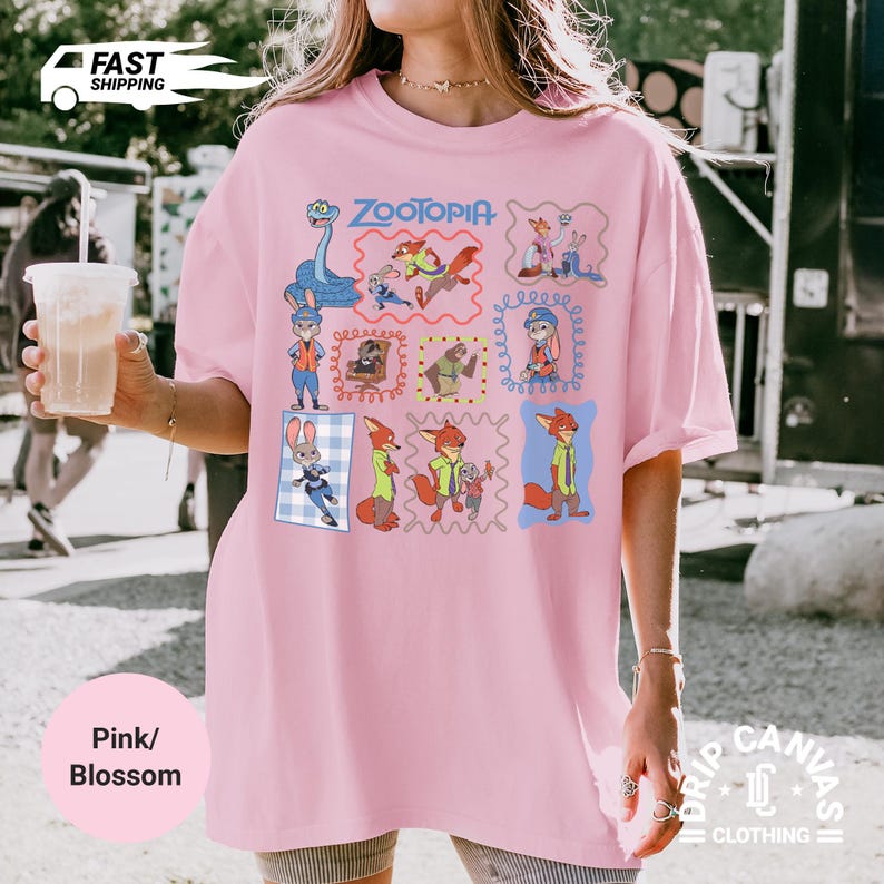 May include: Pink t-shirt with a Zootopia graphic featuring various characters in framed illustrations. The shirt has a crew neck and short sleeves. The text "Zootopia" is in blue script above the character illustrations.