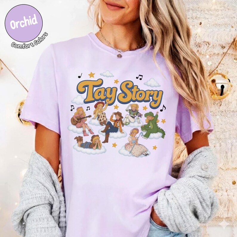 May include: A light purple short-sleeve t-shirt featuring the text Tay Story in yellow with a brown outline. The graphic includes various illustrated characters dressed in cowboy-themed outfits sitting on white clouds with stars and musical notes.