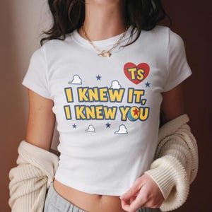 May include: A white short-sleeved t-shirt featuring a graphic print with the text "I KNEW IT, I KNEW YOU" in yellow with blue outlines. Above the text is a red heart containing the letters "TS" and several small blue stars and white clouds.