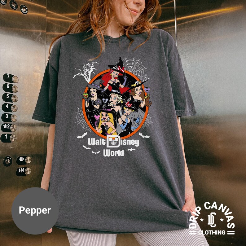 Puede incluir: Camiseta gris oscuro con un dise&ntilde;o de Halloween de princesas de Disney vestidas de brujas, dentro de un c&iacute;rculo naranja. El texto "Walt Disney World" est&aacute; debajo. La camiseta es de color pimienta.