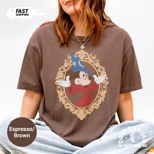 May include: Espresso brown t-shirt with a vintage graphic of Mickey Mouse as a sorcerer, set in a gold oval frame. The shirt has short sleeves and a relaxed fit. The text "Fast Shipping" is in the upper left corner.