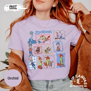 May include: A light purple t-shirt featuring a colorful graphic of characters from the movie "Zootopia". The design includes various framed illustrations of the characters. The shirt also has the word "Zootopia" printed on it.