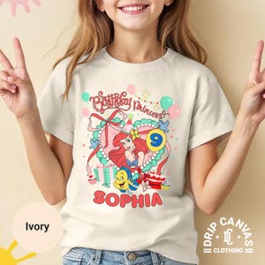 May include: Ivory-colored t-shirt featuring a colorful graphic of a princess with red hair, a blue top, and the text "Birthday Princess" and "Sophia". The number 9 is also displayed. The shirt has short sleeves.
