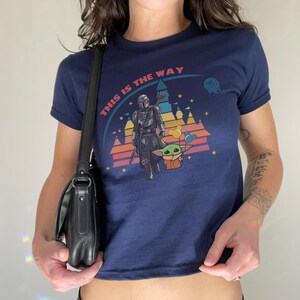May include: Navy blue t-shirt featuring a graphic of The Mandalorian and Grogu with the text "THIS IS THE WAY" in a rainbow arc. The design includes a castle and stars. The shirt has short sleeves.