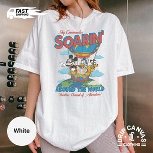 May include: White t-shirt featuring a colorful graphic of Mickey Mouse, Donald Duck, and Goofy in a hot air balloon with the text "SOARIN'" and "AROUND THE WORLD". The shirt has a crew neck and short sleeves.