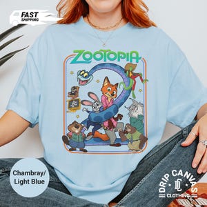 May include: Light blue t-shirt featuring the word "ZOOTOPIA" in green and orange, with cartoon characters from the movie. The shirt has a round neck and short sleeves. The text "Chambray/Light Blue" is printed on the bottom left.