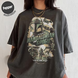 Pu&ograve; includere: T-shirt grigio scuro con grafica della serie The Mandalorian. Il design include il casco Mandaloriano, Grogu e il testo "The Mandalorian and Grogu" e "This is the Way". La maglietta &egrave; etichettata "Pepper Comfort Colors".