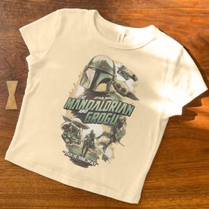 Pu&ograve; includere: T-shirt color crema con un design grafico della serie Star Wars, "The Mandalorian and Grogu". Il design include il casco Mandaloriano, Grogu e la frase "This is the Way".