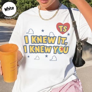 May include: A white t-shirt featuring the yellow text "I KNEW IT, I KNEW YOU" with a blue outline. The design includes a red heart with "TS" in yellow, small blue stars, and white cloud outlines. A colorful ball icon replaces the letter O.
