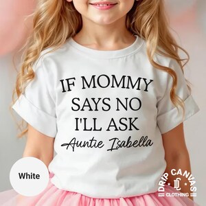 May include: White t-shirt with the text "IF MOMMY SAYS NO I'LL ASK Auntie Isabella" in black. The shirt is worn by a child with blonde hair and a pink tutu. The shirt has rolled-up sleeves.