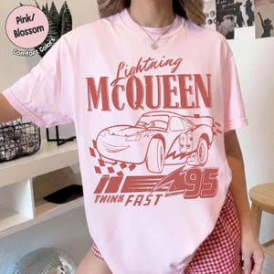 K&ouml;nnte beinhalten: Hellrosa T-Shirt mit einem Lightning McQueen-Motiv aus dem Film Cars. Das Design beinhaltet den Text "Lightning McQueen", eine Rennwagen-Illustration und die Worte "Think Fast" mit der Nummer "95". Das Shirt ist mit "Pink/Blossom" und "Comfort Colors" gekennzeichnet.
