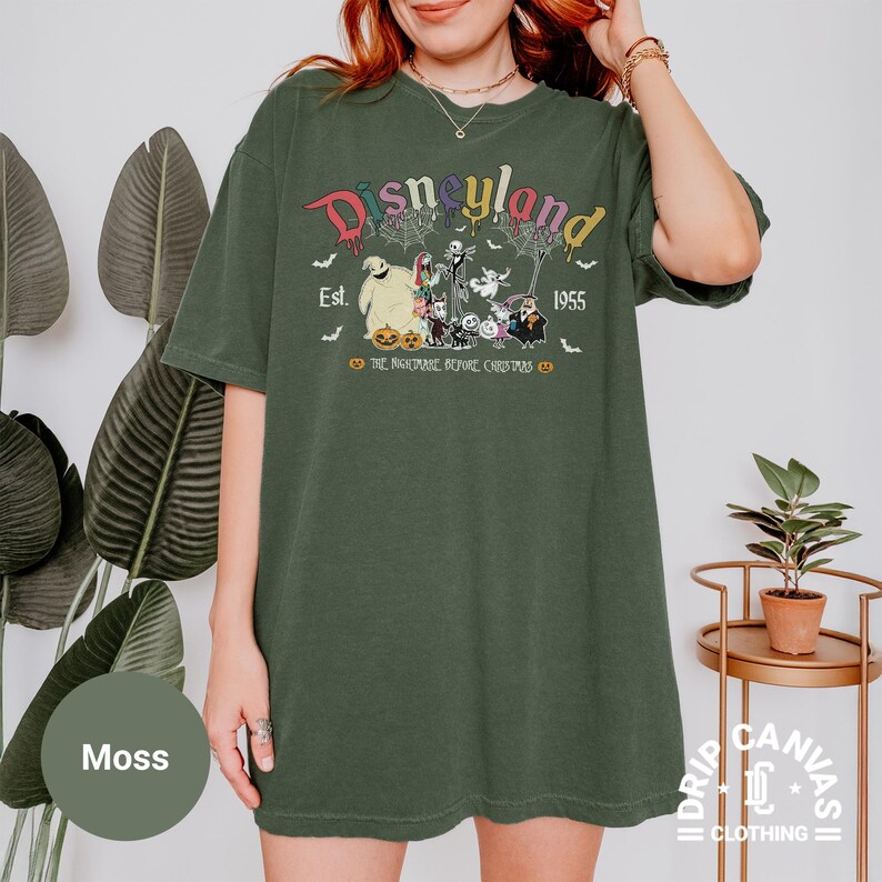 May include: Moss green t-shirt featuring a Disneyland graphic with characters from The Nightmare Before Christmas. The design includes the text "Disneyland Est. 1955" and character illustrations. The shirt is a relaxed fit.