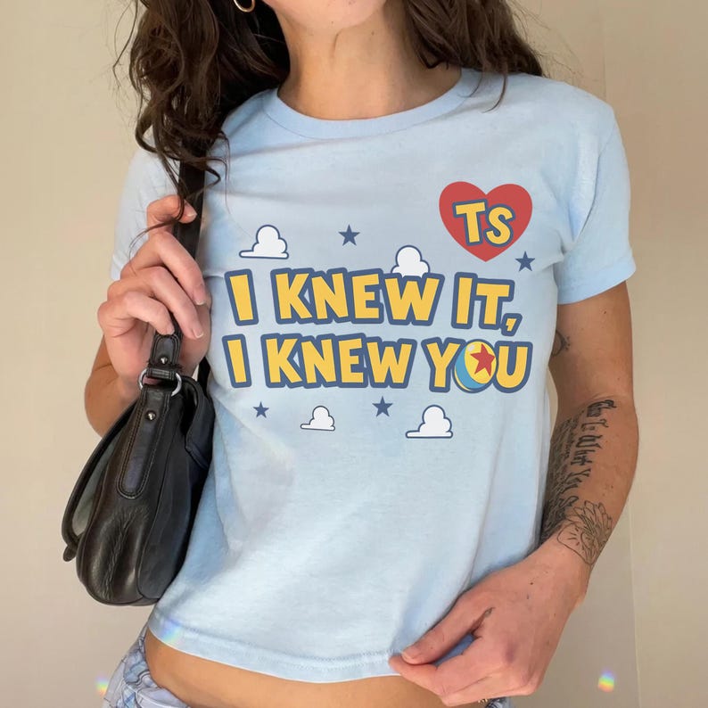 May include: A light blue short-sleeve t-shirt featuring a red heart with the letters TS, yellow text reading I KNEW IT, I KNEW YOU, and a graphic of a ball with a red star, surrounded by small white clouds and dark blue stars on the front.
