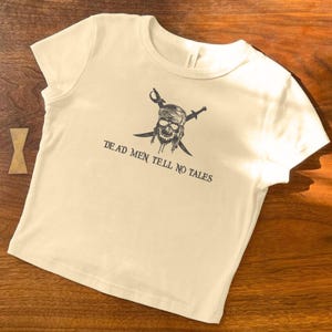 May include: Cream-colored t-shirt featuring a pirate skull graphic with crossed swords and the text "DEAD MEN TELL NO TALES" in black. The shirt has short sleeves and a crew neck.