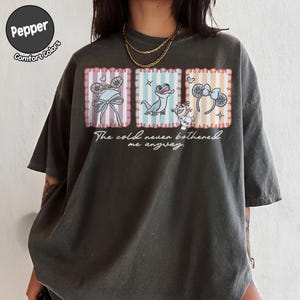 May include: Dark grey t-shirt with three framed illustrations of Disney characters and the text "The cold never bothered me anyway." The illustrations include a bow, a lizard, and a snowman.