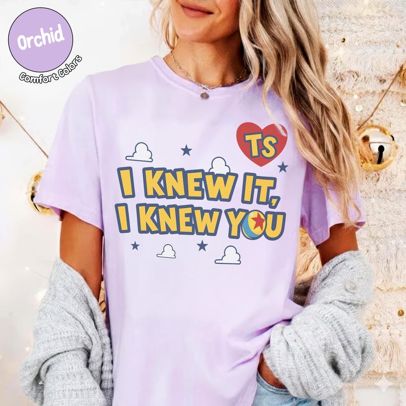 May include: A light purple short-sleeve t-shirt featuring the text I KNEW IT, I KNEW YOU in yellow with blue outlines. Above the text is a red heart with the letters TS and a small ball graphic. The design includes small blue stars and cloud outlines.