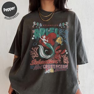 May include: A charcoal gray short-sleeve t-shirt featuring a graphic print of Ariel, Flounder, and Sebastian. The text reads Atlantica Ariel Ocean Tour 1991, Sebastian with the Hot Crustacean Band. A circular logo in the corner says Pepper Comfort Colors.