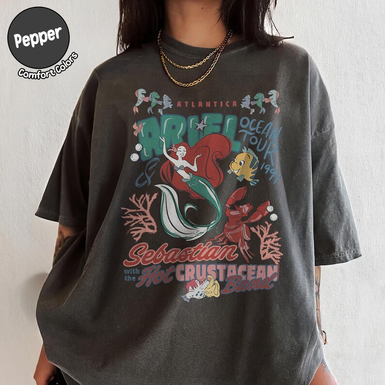 May include: A charcoal gray short-sleeve t-shirt featuring a graphic print of Ariel, Flounder, and Sebastian. The text reads Atlantica Ariel Ocean Tour 1991, Sebastian with the Hot Crustacean Band. A circular logo in the corner says Pepper Comfort Colors.