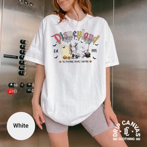 May include: White t-shirt featuring a colorful "Disneyland" graphic with "Est. 1955" and characters from "The Nightmare Before Christmas." The design includes bats, pumpkins, and spiderwebs. The shirt is from "Drip Canvas Clothing."