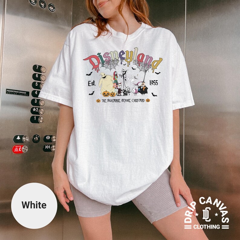 May include: White t-shirt featuring a colorful "Disneyland" graphic with "Est. 1955" and characters from "The Nightmare Before Christmas." The design includes bats, pumpkins, and spiderwebs. The shirt is from "Drip Canvas Clothing."