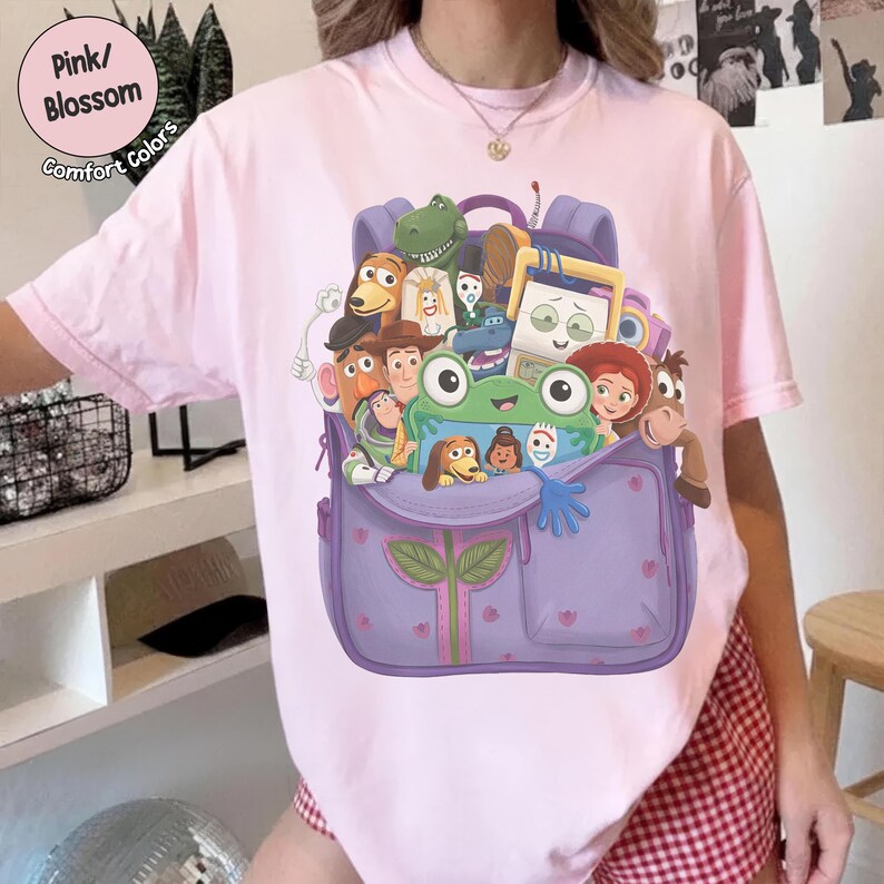 Puede incluir: Una camiseta de manga corta de color rosa claro con un estampado gr&aacute;fico de una mochila morada llena de varios personajes de dibujos animados, incluyendo una rana verde, un vaquero y un ranger espacial. La camiseta tiene cuello redondo.