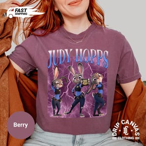 May include: Berry-colored t-shirt featuring the text "JUDY HOPPS" above a graphic of three cartoon bunnies in police uniforms. The graphic has a blue lightning bolt background. The shirt has a crew neck and short sleeves.