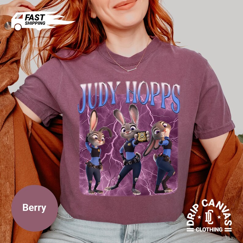 May include: Berry-colored t-shirt featuring the text "JUDY HOPPS" above a graphic of three cartoon bunnies in police uniforms. The graphic has a blue lightning bolt background. The shirt has a crew neck and short sleeves.