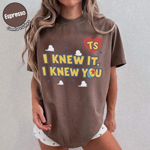 May include: A brown short-sleeve t-shirt featuring a red heart with the letters TS, the text I KNEW IT, I KNEW YOU in yellow, and small white cloud and blue star graphics. The shirt has a crew neckline and a relaxed fit with a casual appearance.