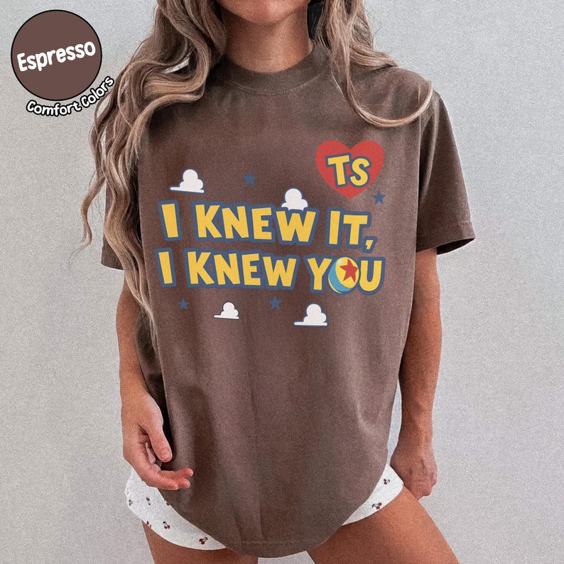 May include: A brown short-sleeve t-shirt featuring a red heart with the letters TS, the text I KNEW IT, I KNEW YOU in yellow, and small white cloud and blue star graphics. The shirt has a crew neckline and a relaxed fit with a casual appearance.