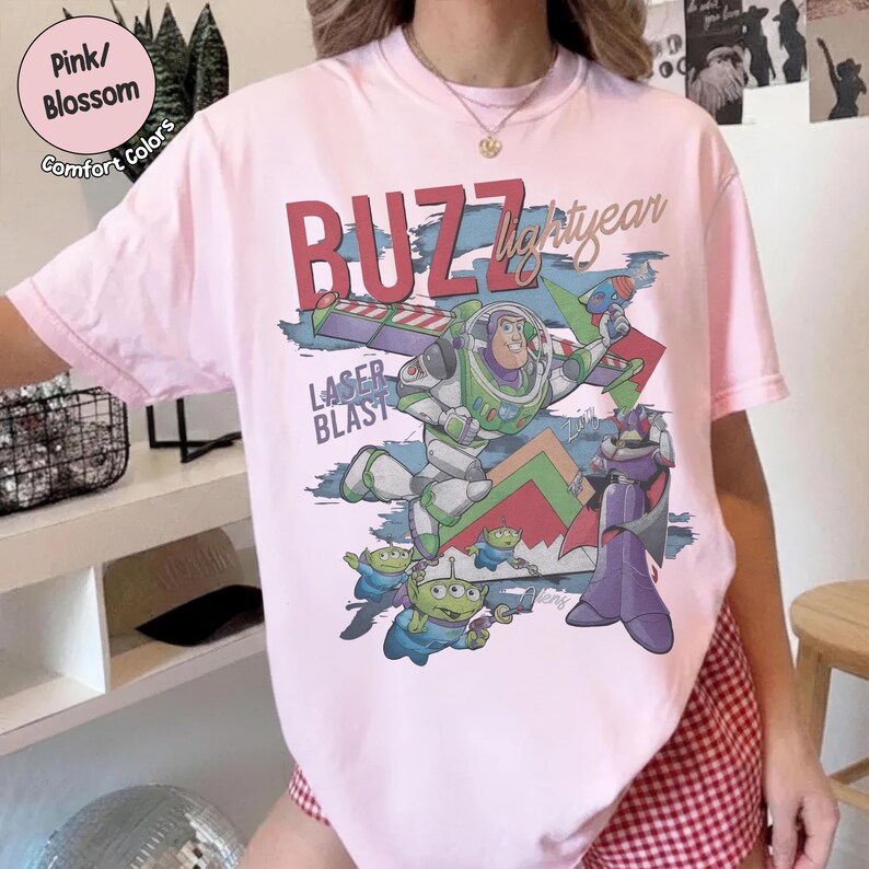 Może przedstawiać: Jasnor&oacute;żowy t-shirt z nadrukiem graficznym przedstawiającym Buzza Astrala, Zurga i kilka zielonych kosmit&oacute;w. Napis głosi BUZZ Lightyear oraz LASER BLAST pogrubioną czerwoną i niebieską czcionką. Wz&oacute;r zawiera geometryczne kształty w kolorze zielonym i czerwonym.