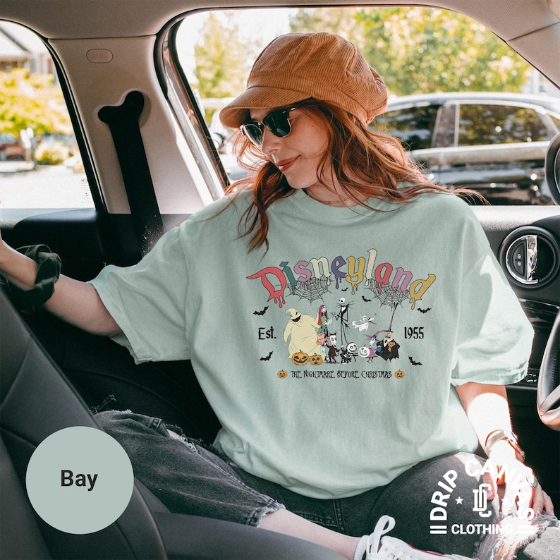 May include: A light green t-shirt with a colorful Disneyland graphic featuring characters from The Nightmare Before Christmas. The design includes the text "Disneyland Est. 1955" and various Halloween-themed elements.