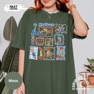 May include: Moss green t-shirt featuring a colorful graphic of characters from the animated film "Zootopia." The design includes the film's title and various character illustrations within decorative frames. The shirt has a relaxed fit and short sleeves.