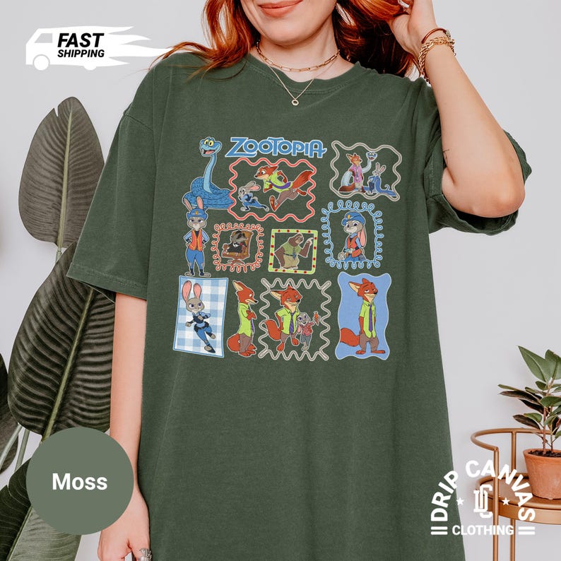 May include: Moss green t-shirt featuring a colorful graphic of characters from the animated film "Zootopia." The design includes the film's title and various character illustrations within decorative frames. The shirt has a relaxed fit and short sleeves.