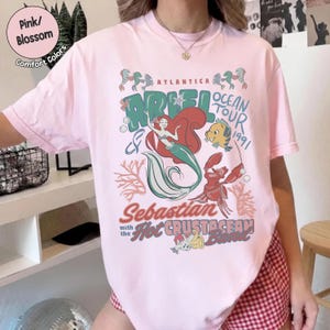 May include: A light pink short-sleeve t-shirt featuring a graphic print of Ariel, Flounder, and Sebastian. The text reads Atlantica Ariel Ocean Tour 1991, Sebastian with the Hot Crustacean Band, and includes illustrations of coral and bubbles.