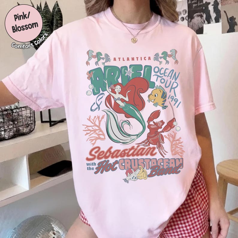 May include: A light pink short-sleeve t-shirt featuring a graphic print of Ariel, Flounder, and Sebastian. The text reads Atlantica Ariel Ocean Tour 1991, Sebastian with the Hot Crustacean Band, and includes illustrations of coral and bubbles.