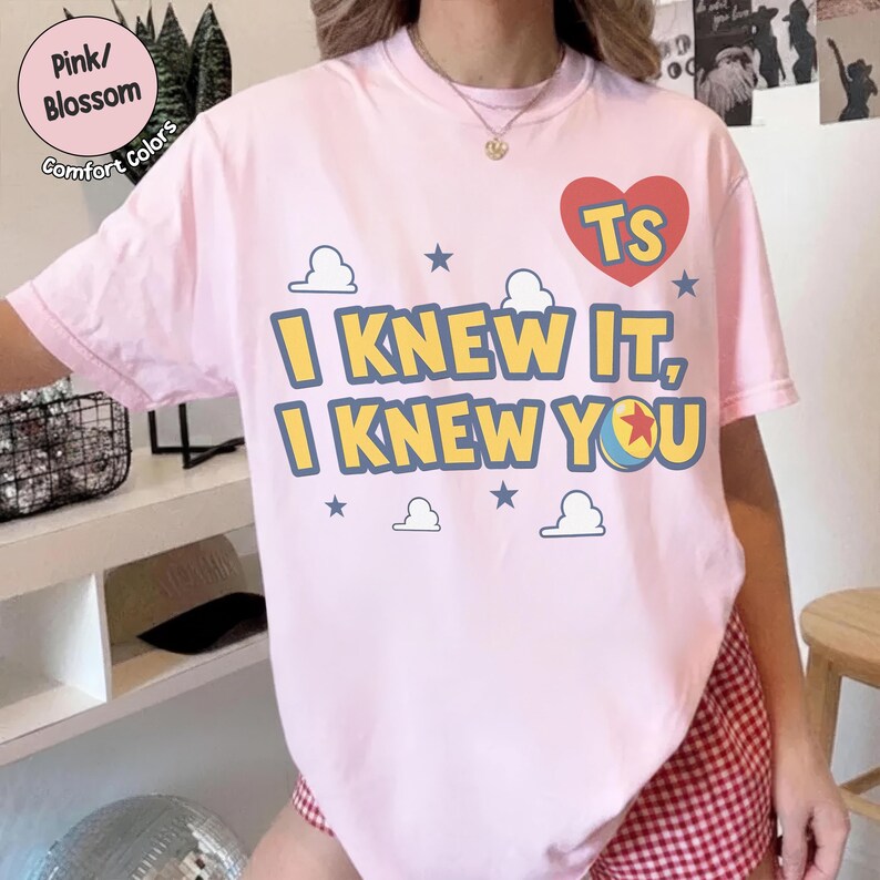 May include: A light pink short-sleeve t-shirt featuring a red heart with the letters TS, the text I KNEW IT, I KNEW YOU in yellow with blue outlines, and a graphic of a ball with a red star, surrounded by small blue stars and white clouds.