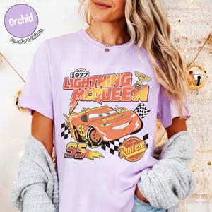 Op de afbeelding: Lichtpaars T-shirt met een Lightning McQueen-afbeelding uit de film Cars. Het ontwerp bevat de tekst "Lightning McQueen" en "95" met een geblokte vlag en een trofee. Het shirt is van Comfort Colors.