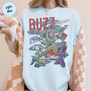 Może przedstawiać: Jasnoniebieska bluza z okrągłym dekoltem z nadrukiem graficznym przedstawiającym Buzza Astrala, Zurga i zielonych kosmit&oacute;w. Napis głosi BUZZ Lightyear oraz LASER BLAST czerwoną i brązową czcionką. Wz&oacute;r zawiera geometryczne kształty i niebieskie pociągnięcia pędzla.