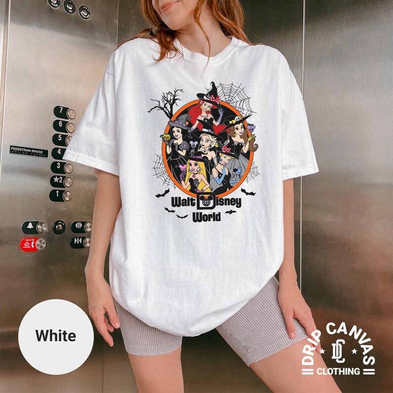 Puede incluir: Camiseta blanca con un dise&ntilde;o circular de princesas de Disney vestidas de brujas, con telara&ntilde;as y murci&eacute;lagos. El texto "Walt Disney World" est&aacute; debajo. Parte de la colecci&oacute;n Drip Canvas Clothing.