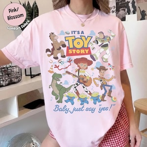 Op de afbeelding: Een lichtroze T-shirt met een kleurrijke Toy Story-afbeelding met personages zoals Woody, Buzz Lightyear, Jessie, Rex, Bullseye, Forky en drie aliens. De tekst luidt IT'S A TOY STORY en Baby, just say 'yes!' in blauw en rood.