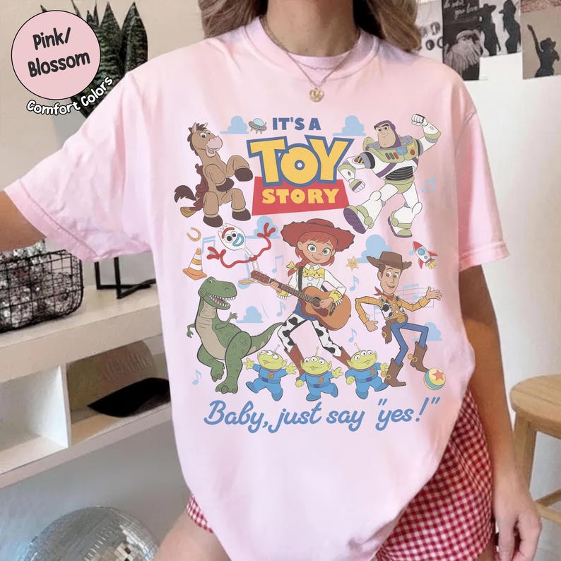 Op de afbeelding: Een lichtroze T-shirt met een kleurrijke Toy Story-afbeelding met personages zoals Woody, Buzz Lightyear, Jessie, Rex, Bullseye, Forky en drie aliens. De tekst luidt IT'S A TOY STORY en Baby, just say 'yes!' in blauw en rood.