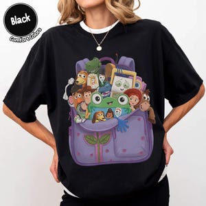 Puede incluir: Una camiseta negra con un estampado gr&aacute;fico de una mochila morada llena de varios personajes de juguetes animados, incluyendo una rana verde, un vaquero, un guardi&aacute;n espacial y una figura con forma de patata en estilo de dibujos.