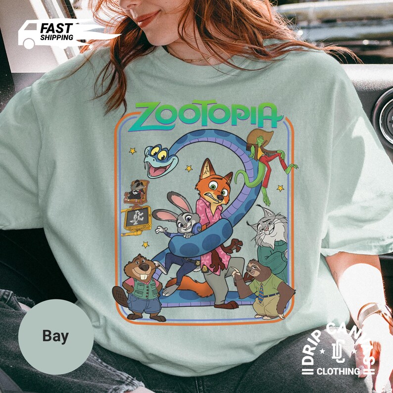 May include: A light blue t-shirt featuring a colorful graphic from the animated movie "Zootopia." The design includes characters like Nick Wilde, Judy Hopps, and others, with the movie title in a retro font. The shirt has a crew neck.