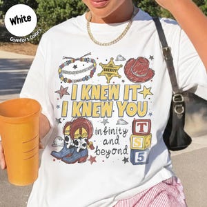 以下が含まれることがあります： I KNEW IT I KNEW YOU infinity and beyondというテキストがプリントされた白いTシャツ。デザインには、保安官バッジ、カウボーイハット、ビーズのブレスレット、牛柄のカウボーイブーツ、T S 5と書かれた3つの積み木が描かれています。