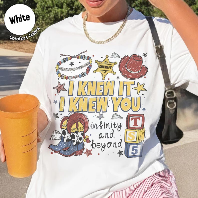 以下が含まれることがあります： I KNEW IT I KNEW YOU infinity and beyondというテキストがプリントされた白いTシャツ。デザインには、保安官バッジ、カウボーイハット、ビーズのブレスレット、牛柄のカウボーイブーツ、T S 5と書かれた3つの積み木が描かれています。