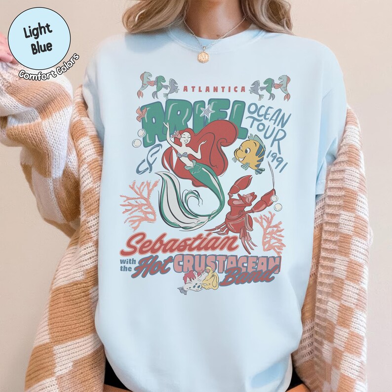 May include: A light blue crewneck sweatshirt featuring a graphic print of Ariel, Flounder, and Sebastian. The text reads Atlantica Ariel Ocean Tour 1991, Sebastian with the Hot Crustacean Band. The design includes coral, bubbles, and musical notes.